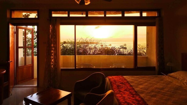A hotel room with a bed, a desk, and a chair. There is a large window with a view of the sea and boats. - Galeodan Pompano Suite (Puerto Baquerizo Moreno)