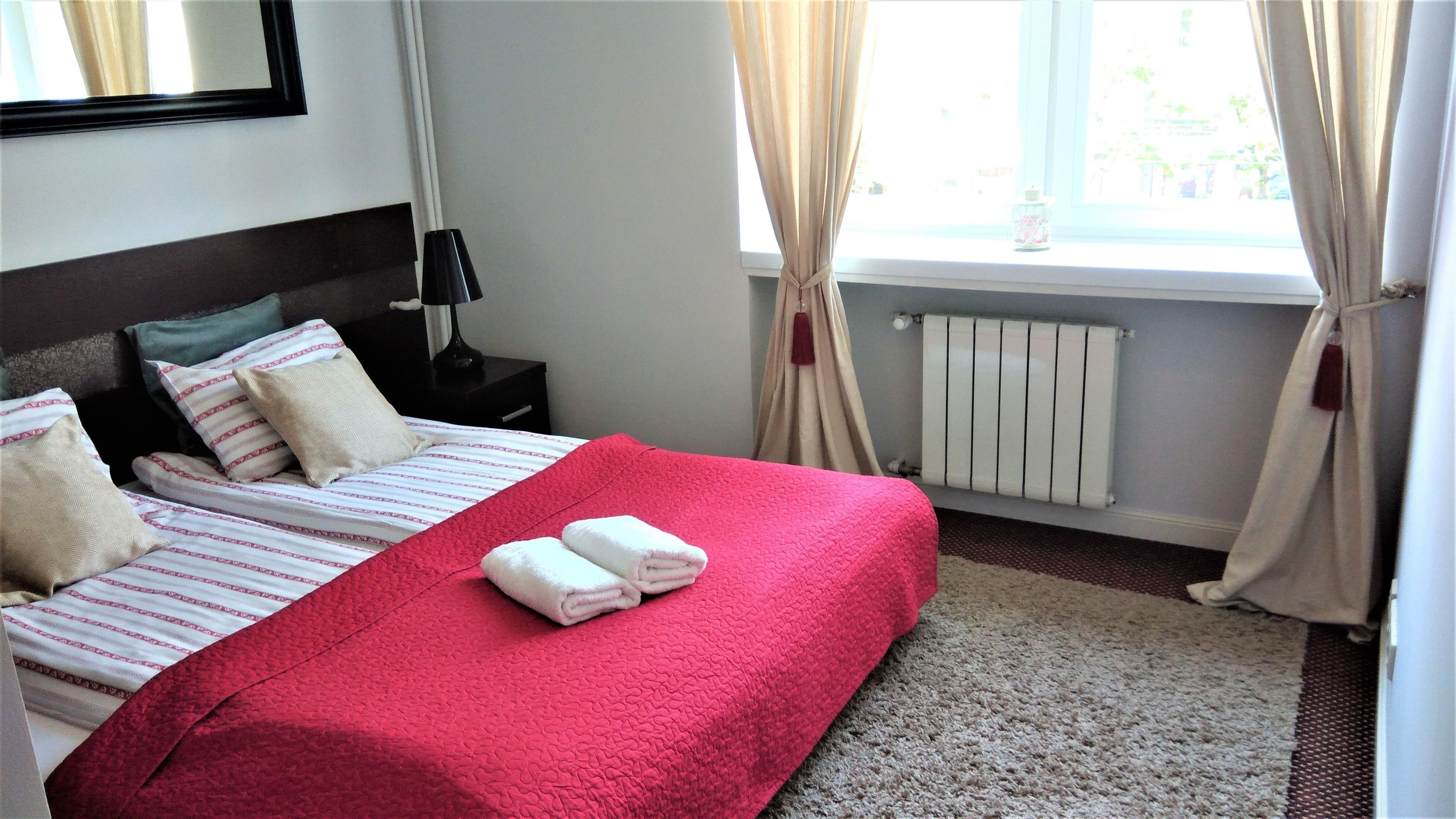A hotel room with two beds, a radiator, a nightstand with a lamp, and a window with curtains.