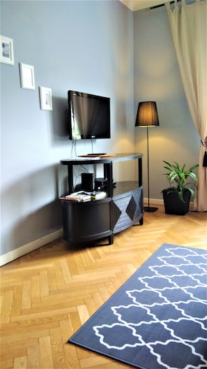 Un salon avec un téléviseur à écran plat fixé au mur, une table d’appoint noire sur laquelle sont posés des appareils électroniques, une lampe sur pied, une plante en pot et un tapis à motifs.
