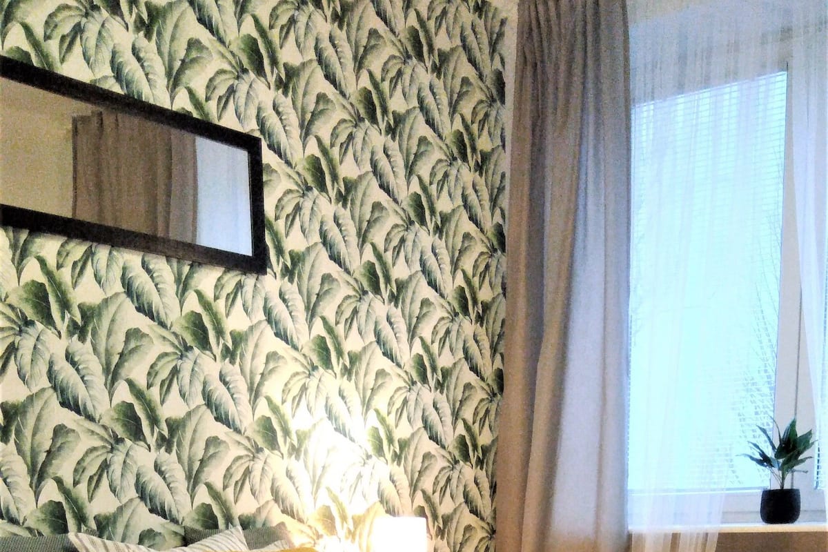 A bedroom with a bed, a nightstand, a lamp, a window with curtains, and a plant.