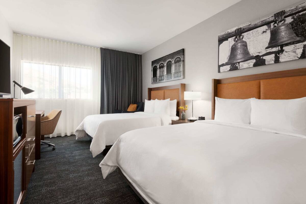 A hotel room with two beds, a desk, a chair, a lamp, and a painting on the wall.