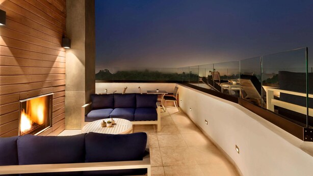 A modern outdoor lounge area with a fireplace, seating, and a glass railing overlooking a cityscape.