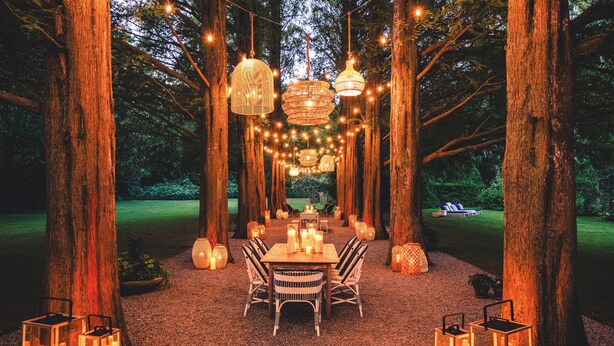 Un área de comedor al aire libre con una mesa y sillas, rodeada de árboles altos y luces colgantes.