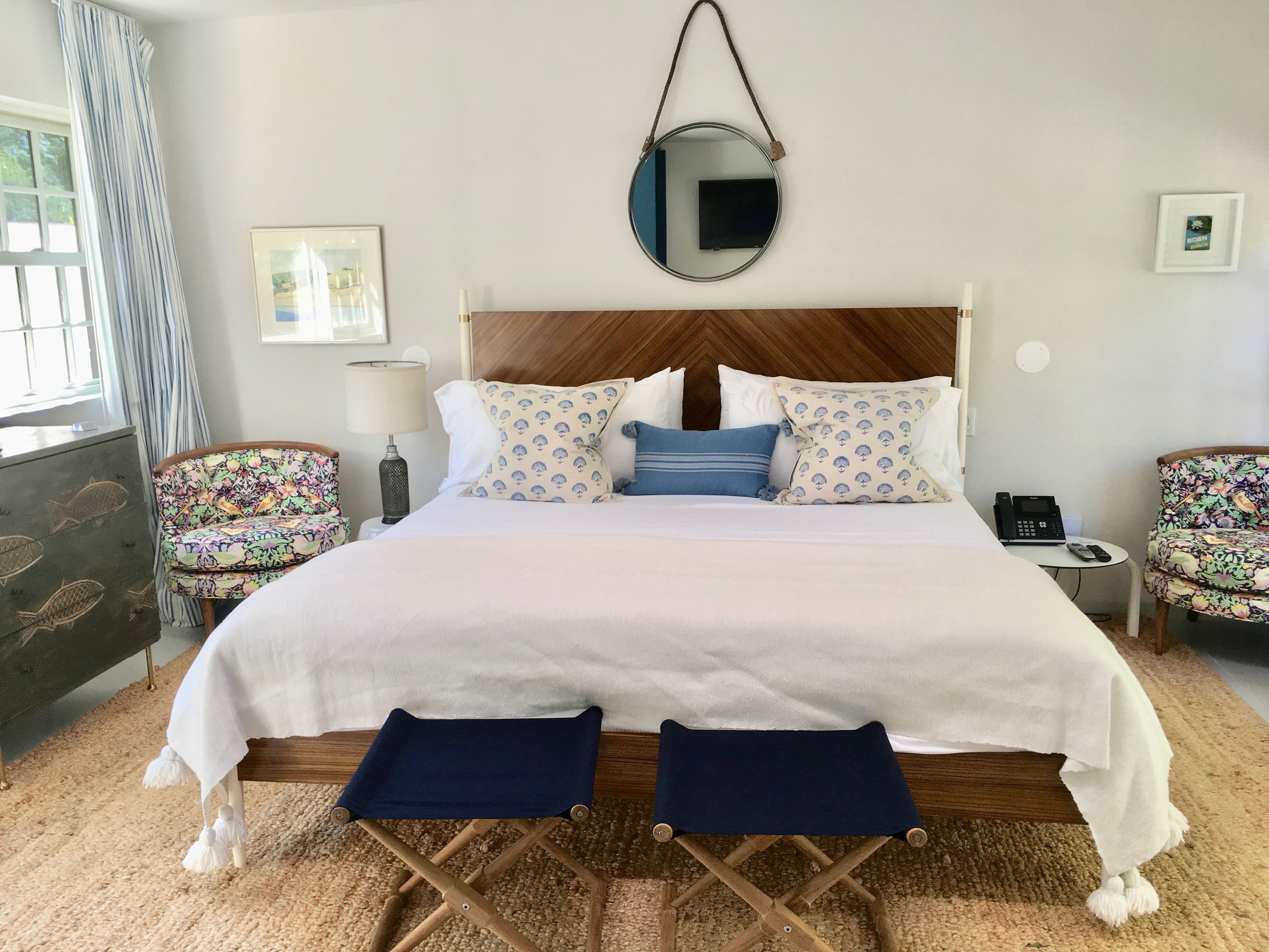 Habitación de hotel ordenada con una cama grande, dos mesitas de noche, un espejo redondo pequeño y una silla estampada con motivos florales.