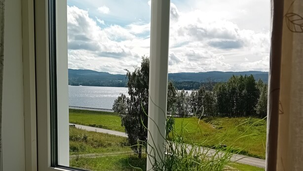 A window with a view of a lake, greenery, and distant hills. - Täljegården Lantgårdshotell (Erikslund)