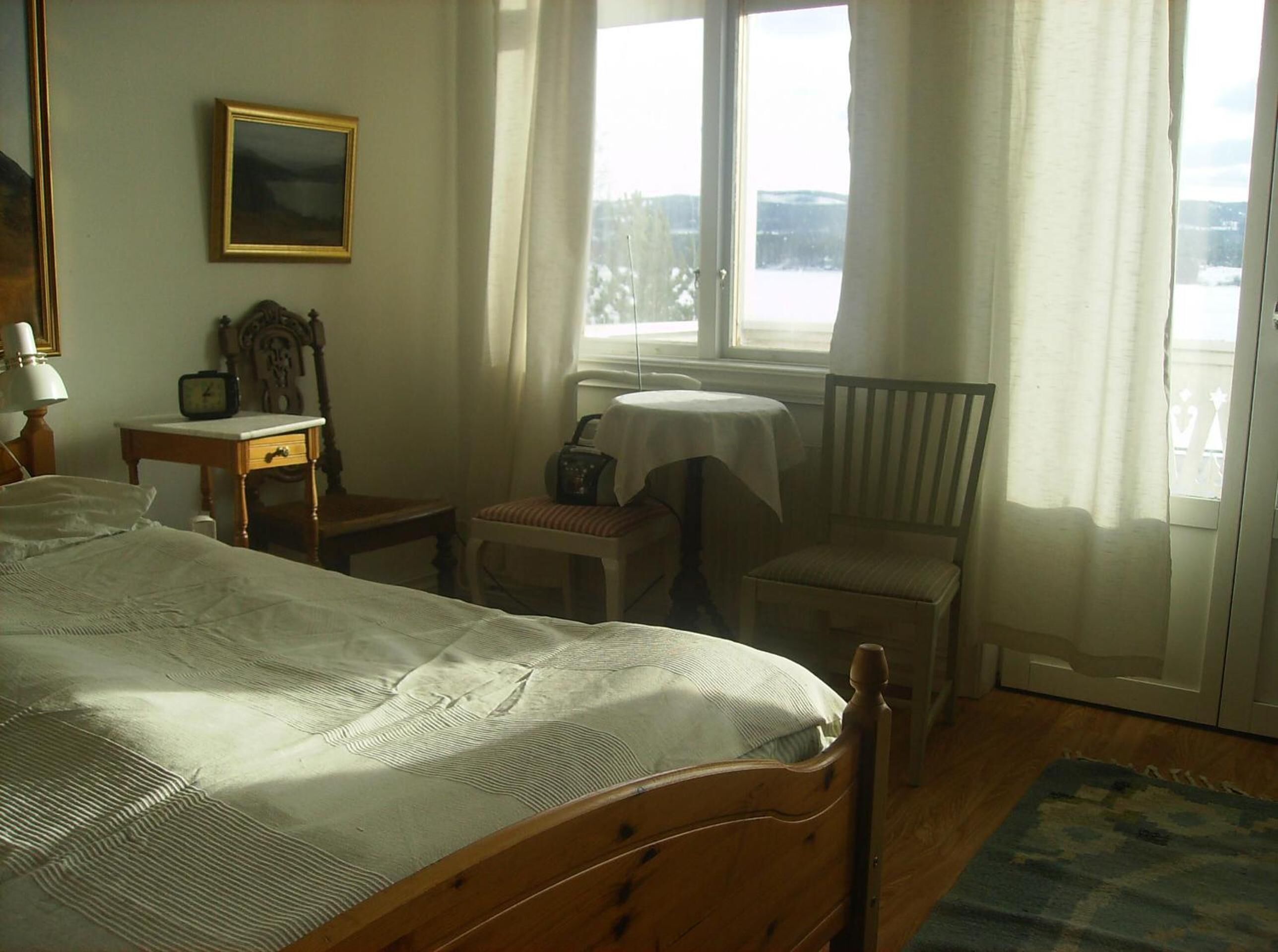 Un dormitorio con una cama, una mesita de noche con lámpara, una silla, una pequeña mesa con radio y una ventana con cortinas.