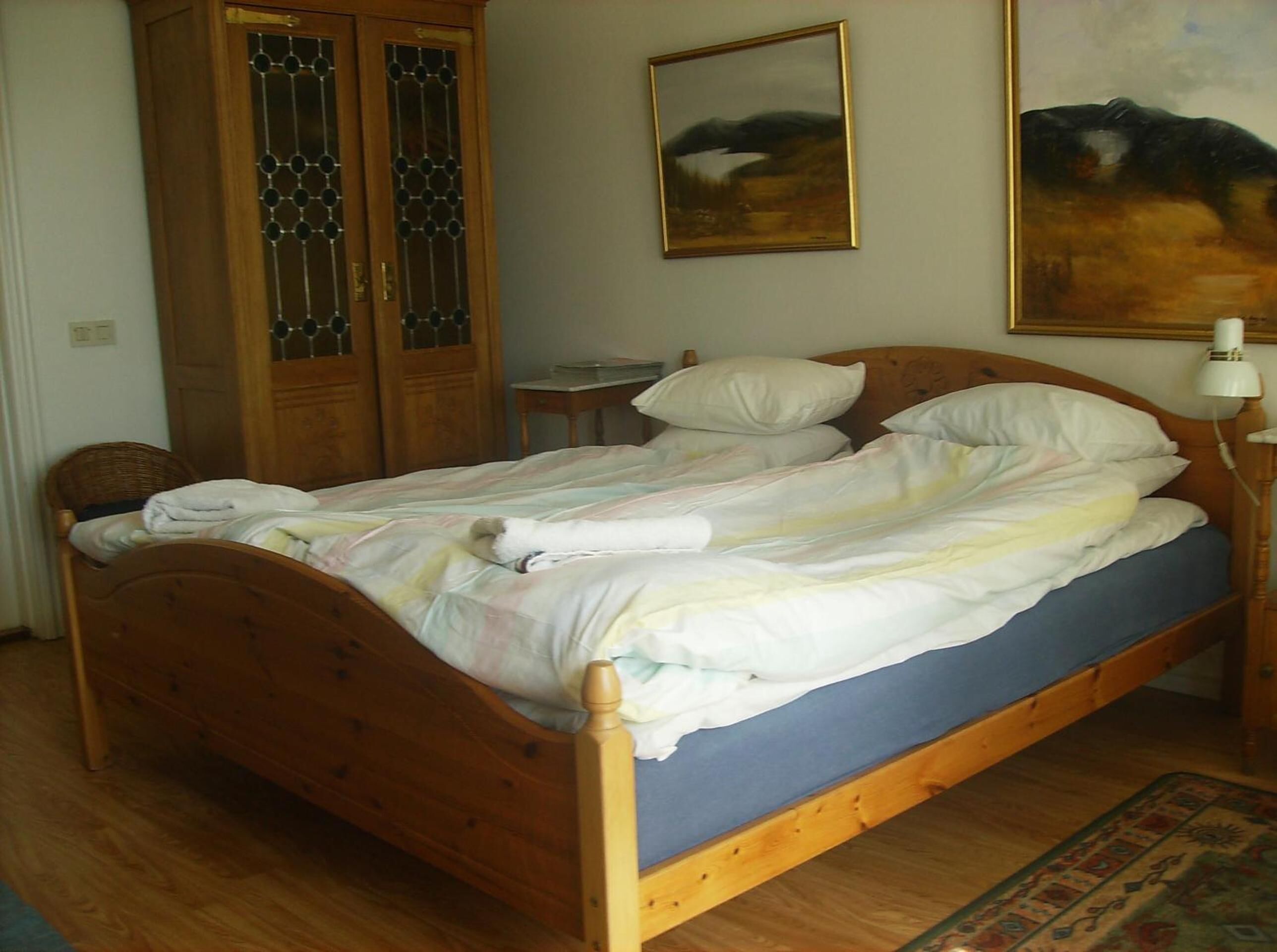 Uma cama de madeira com colchão azul e roupa de cama branca, um guarda-roupa de madeira com portas de vidro, uma pequena mesa de cabeceira e um quadro na parede.