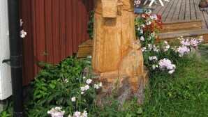 A wooden sculpture of a woman holding a book, placed in front of a red house with white shutters and a garden with pink flowers. - Täljegården Lantgårdshotell (Erikslund)
