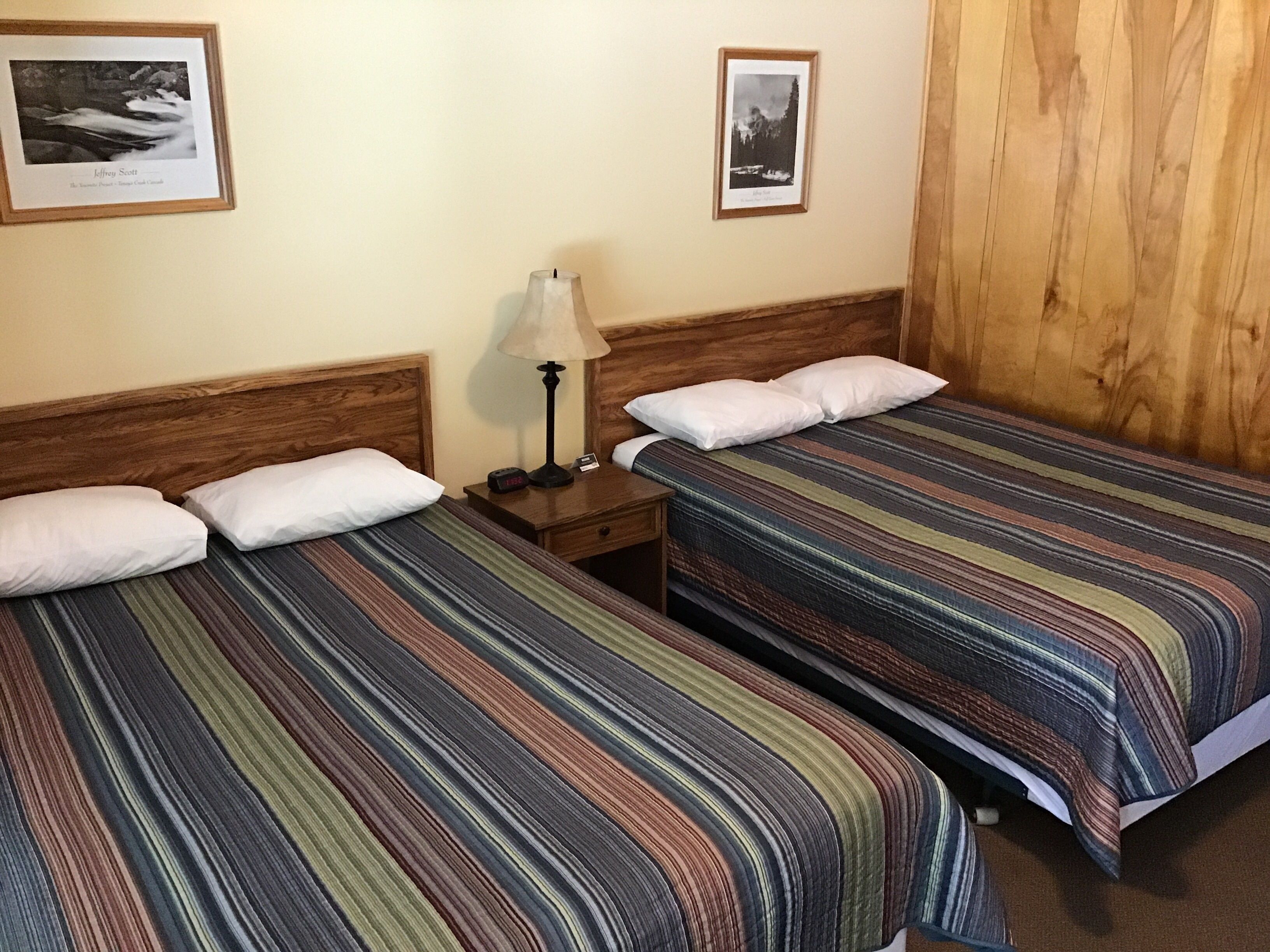 Une chambre d’hôtel avec deux lits simples, des têtes de lit en bois, une table de chevet avec une lampe et des tableaux encadrés accrochés au mur.