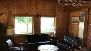 Een houten interieur met een bank, twee stoelen, een ronde tafel, een lamp, een opgeplakt dierenhoofd en een lijst met een afbeelding.