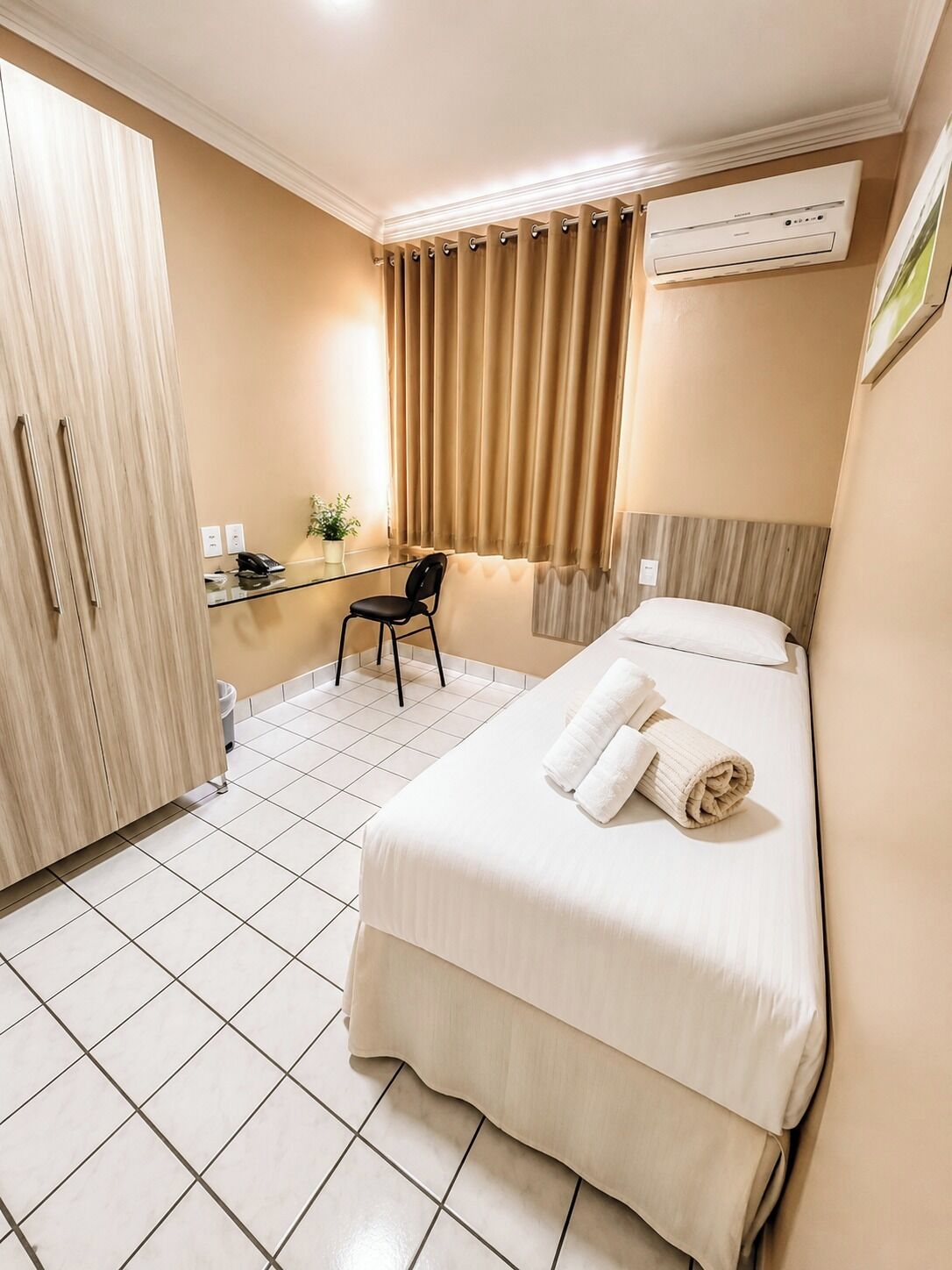 A hotel room with a bed, a desk, a chair, a wardrobe, and an air conditioning unit.