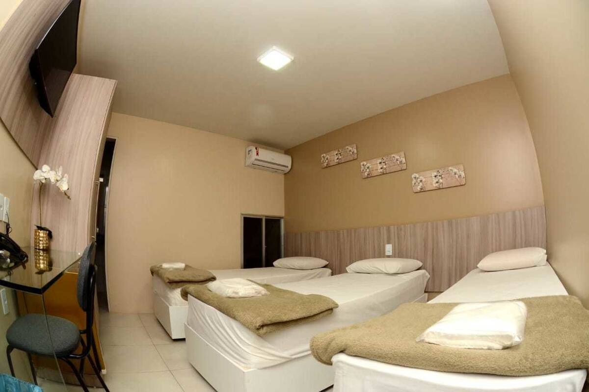 A hotel room with three beds, a desk with a chair, an air conditioning unit, and a wall-mounted TV.