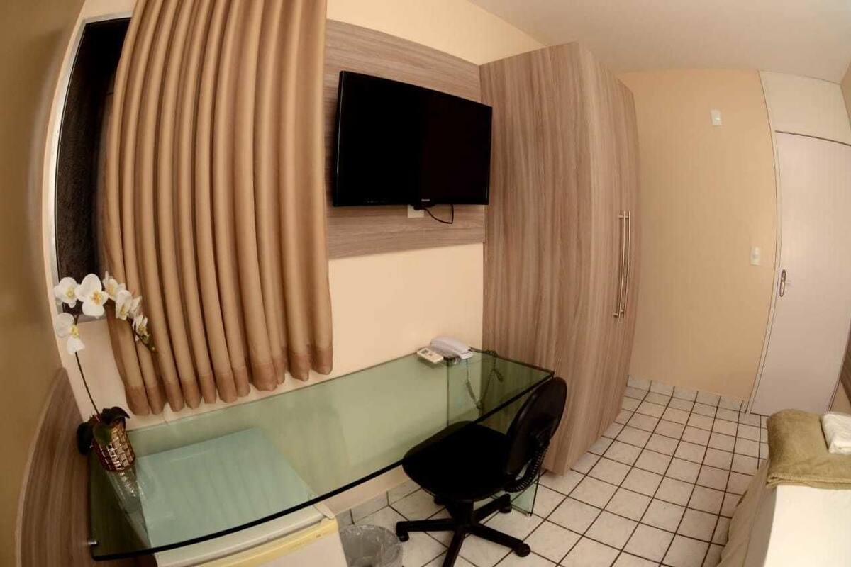 A compact room with a glass desk, a black office chair, a mounted TV, and a wardrobe.