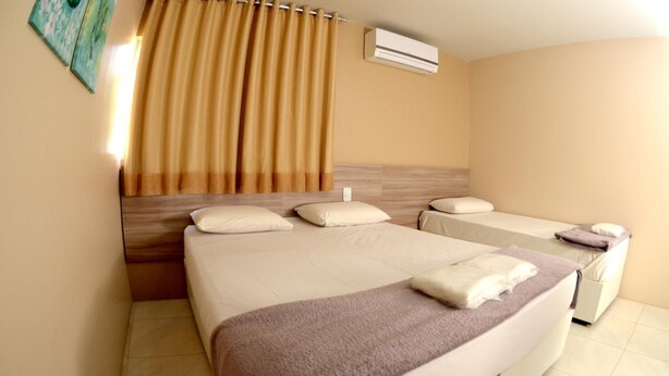 A hotel room with two beds, a large window with curtains, an air conditioning unit, and a wall-mounted artwork. - Hotel Itaparica (Caruaru)