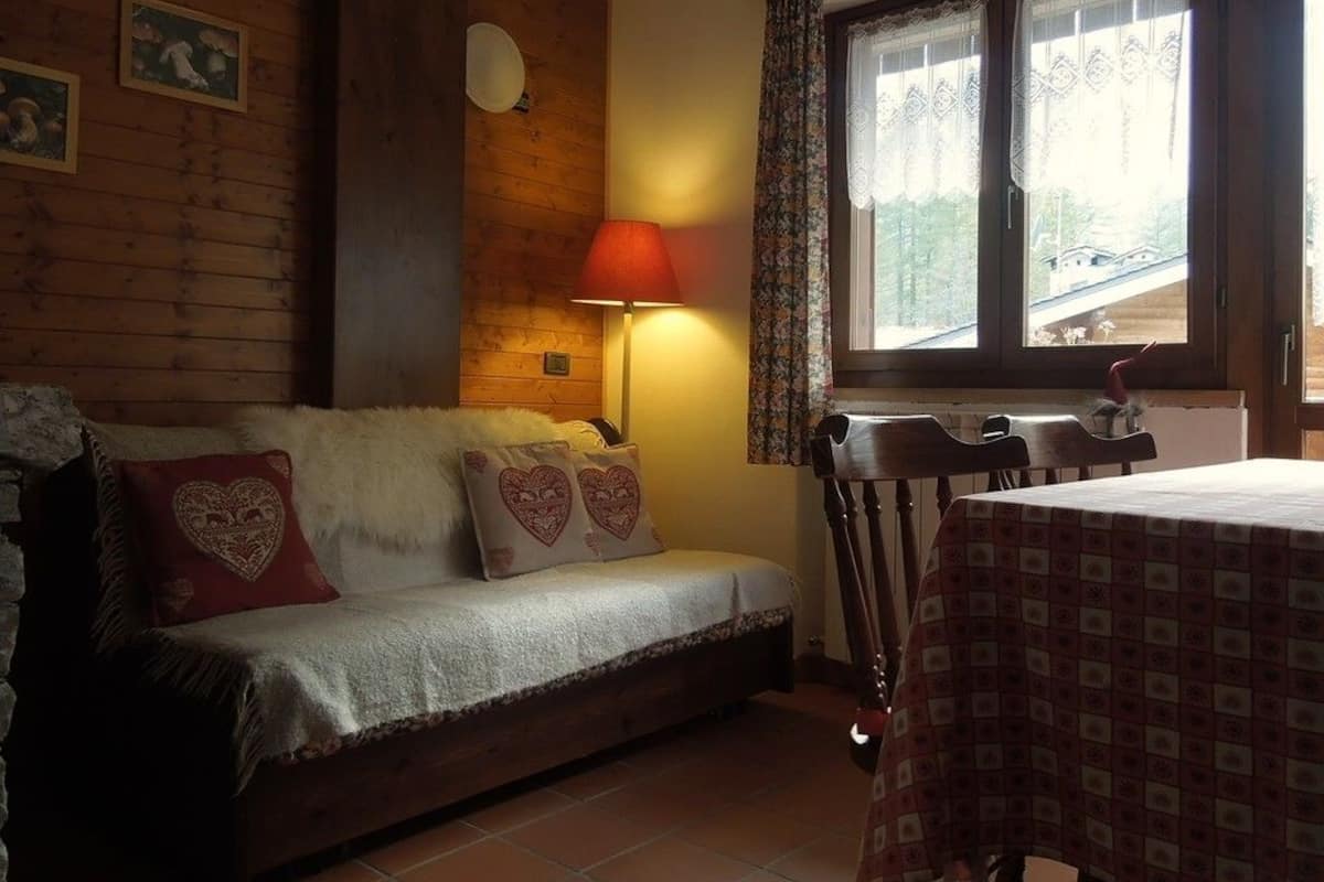 A cozy room with a wooden wall, a sofa with heart-patterned cushions, a table with a patterned tablecloth, and a window with lace curtains.