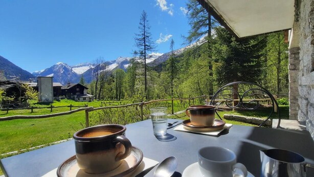 A table with two cups of coffee and a glass of water, set against a backdrop of a mountainous landscape with trees and a wooden building. - Residence Weissthor (Macugnaga)