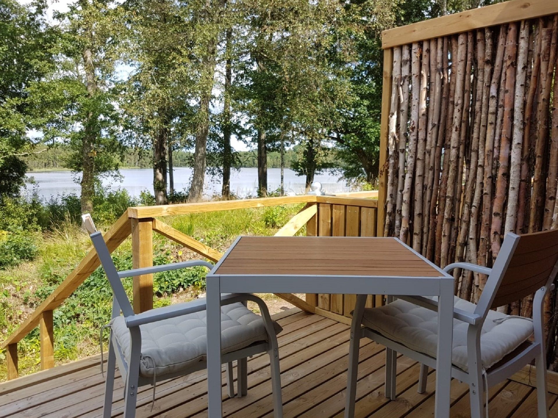 A wooden deck with a table and two chairs, overlooking a lake with trees in the background.