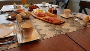 A breakfast table set with plates, bowls, a cutting board with sliced meats, cheese, and bread, along with cups and a vase with flowers. - Den Sovande Älgen (Markaryd)