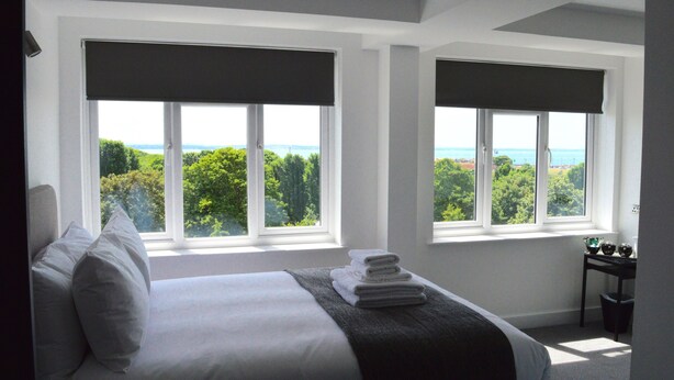 A bedroom with a large bed, two windows with blinds, a view of greenery and the sea, and a small table with a trash bin. - Q8 Boutique Hotel (Portsmouth)