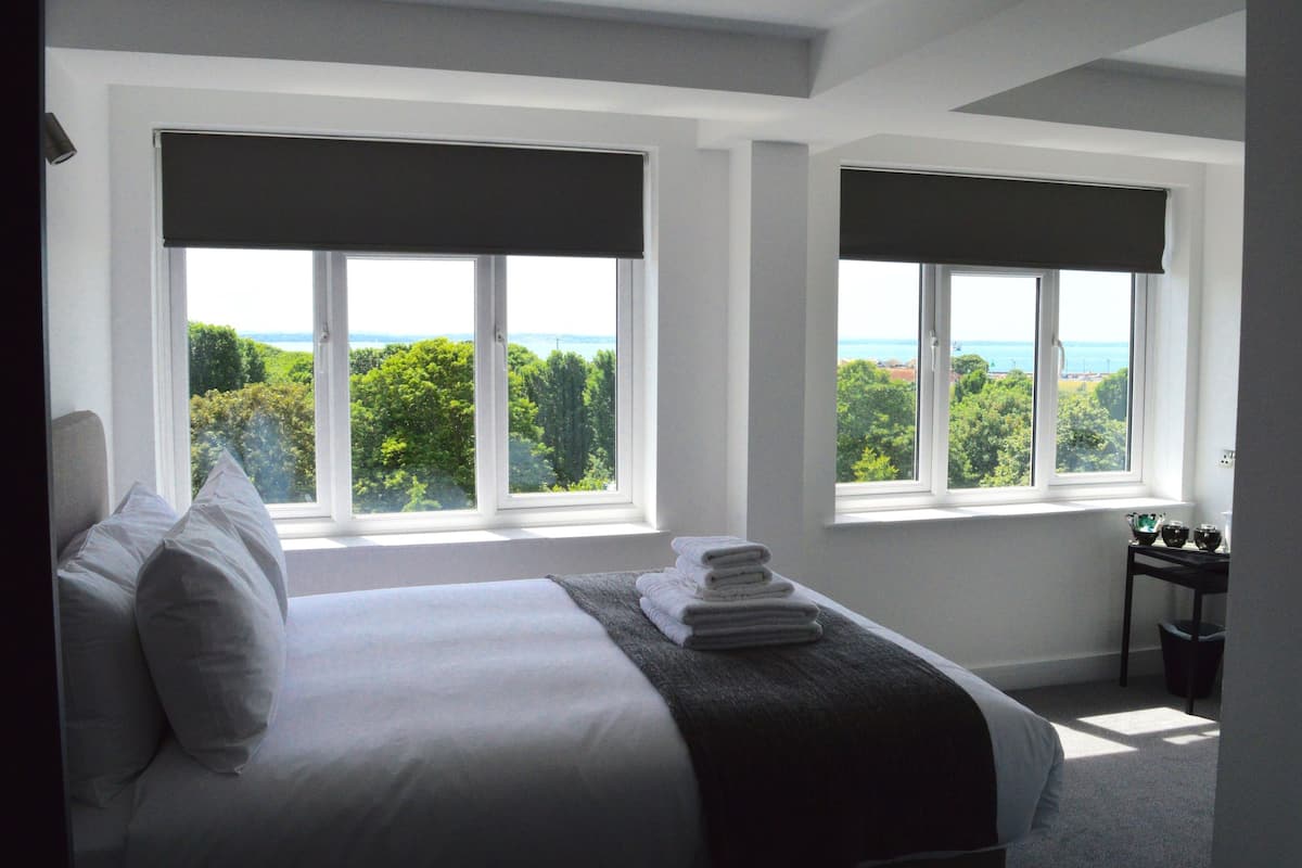 Une chambre avec un grand lit, deux fenêtres équipées de stores, une vue sur la verdure et la mer, et une petite table avec une poubelle.