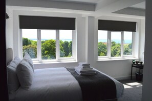 Ein Schlafzimmer mit einem großen Bett, zwei Fenstern mit Jalousien, Blick auf Grünflächen und das Meer sowie einem kleinen Tisch mit einem Mülleimer.