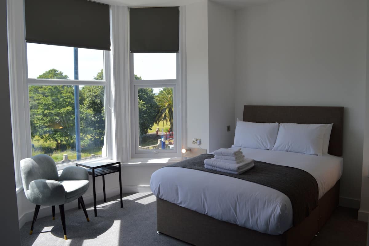 Une chambre à coucher avec un lit, une chaise, une table et une fenêtre donnant sur des arbres et un immeuble.