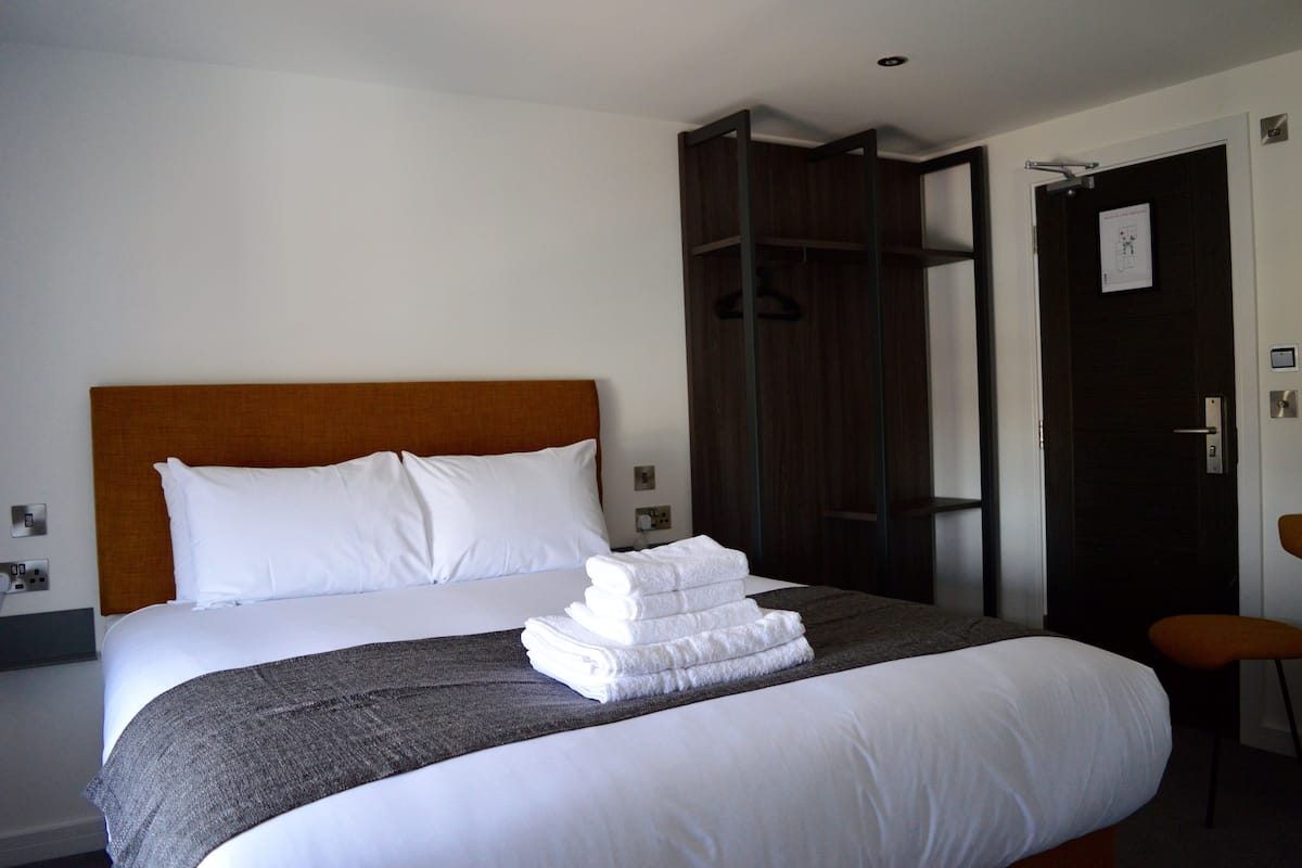Un lit bien fait, avec du linge de lit blanc et une pile de serviettes pliées, une tête de lit en bois et une armoire en bois foncé.