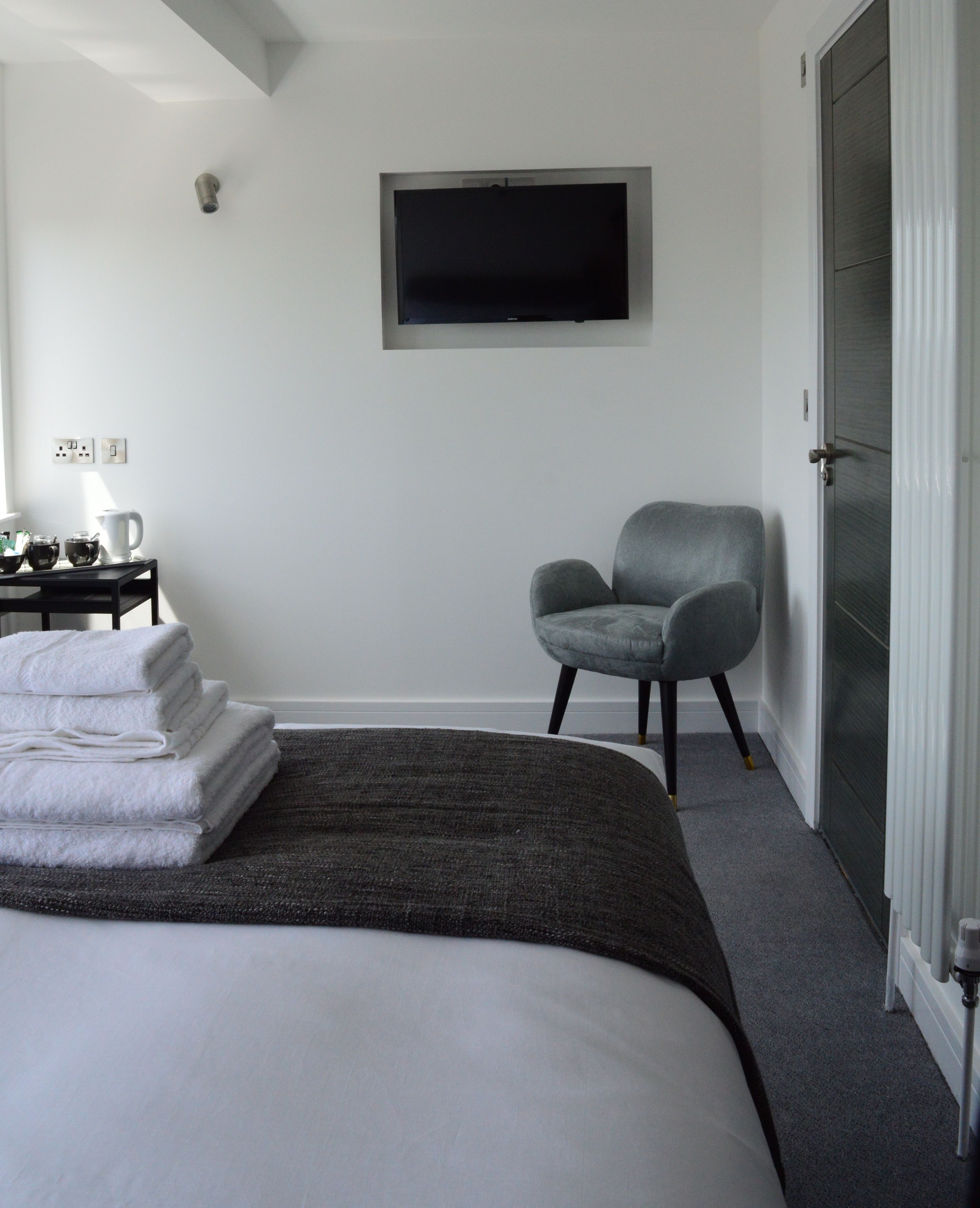 A modern hotel room with a large bed, a grey armchair, a flat-screen TV mounted on the wall, and a door with a glass panel.