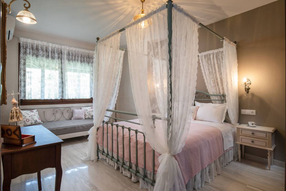 A four-poster canopy bed with sheer curtains, a bedside table, a nightstand, a window with lace curtains, and a sofa with cushions.