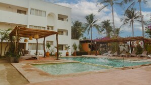 Uma área de piscina com espreguiçadeiras e um pergolado, rodeada por palmeiras e um edifício branco.