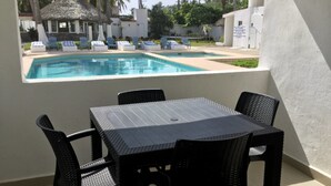 Área junto à piscina com mesa e cadeiras, uma estrutura com telhado de palha e vista para a piscina e palmeiras.