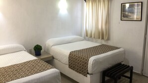 Quarto de hotel com duas camas de solteiro, uma pequena mesa de cabeceira com uma planta, um banco, um ar condicionado, uma janela com cortinas e um quadro na parede.