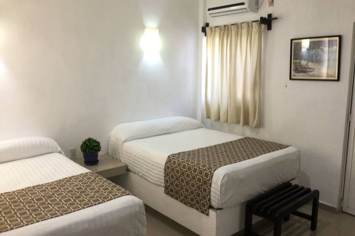 Een hotelkamer met twee bedden, een kleine kamerplant, een airconditioningapparaat, een raam met gordijnen en een lijst met een foto aan de muur.