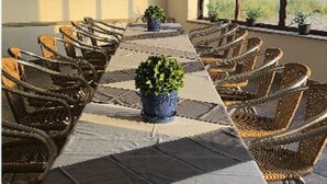 Uma longa mesa de jantar com cadeiras de vime, plantas em vasos e vista para a praia através de grandes janelas.