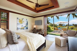 A bedroom with a bed, a desk, and a TV. There is a view of a garden and palm trees outside.