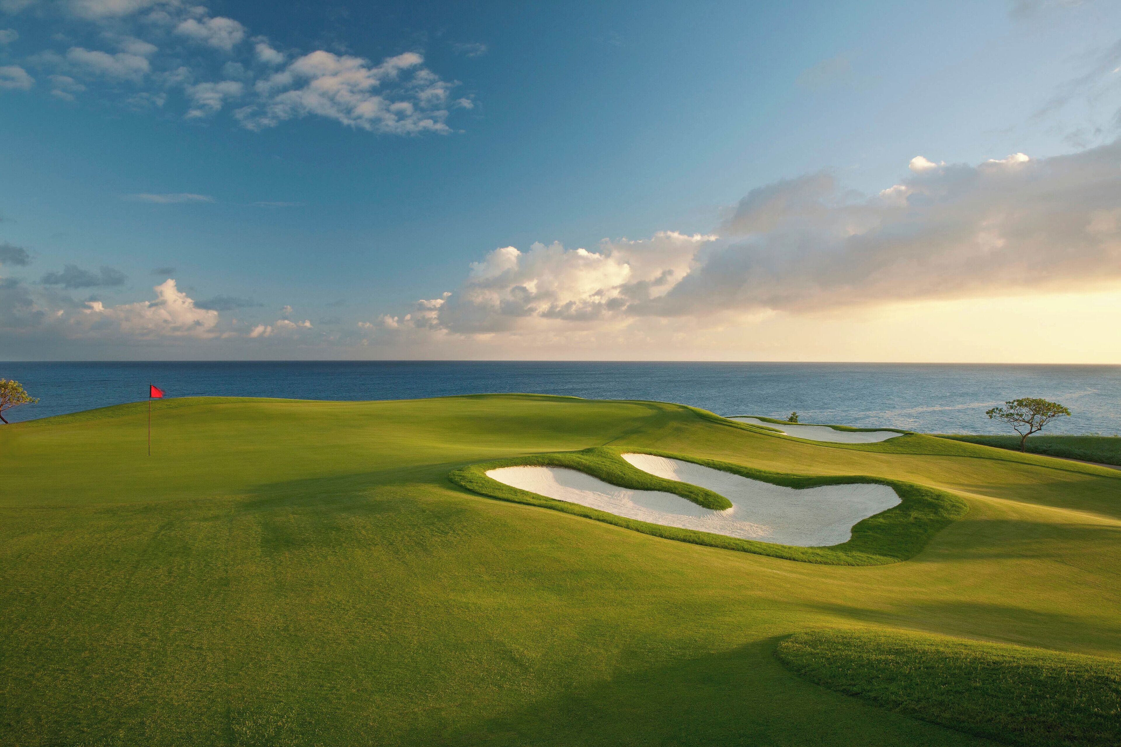 Um campo de golfe com uma bandeira, armadilhas de areia e vista para o oceano.