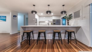 Una cocina moderna con una isla central, taburetes de bar, luces colgantes y vistas al salón contiguo.