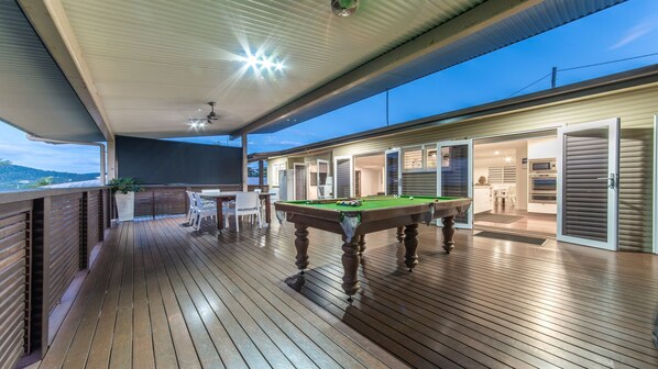 Um deck de madeira com mesa de bilhar e cadeiras, área coberta com ventilador de teto e vista para um edifício residencial com portas deslizantes.