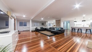 Amplia sala de estar con sofá de cuero negro, piso de madera y cocina moderna con armarios blancos y taburetes de bar.