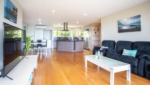 A modern living room with a large flat-screen TV, a glass coffee table, a black sofa, and a kitchen in the background. - Getaway Ocean Views (Cannonvale)