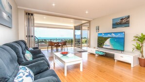 A modern living room with a sofa, a coffee table, a TV, and a balcony with a view of the sea. - Getaway Ocean Views (Cannonvale)