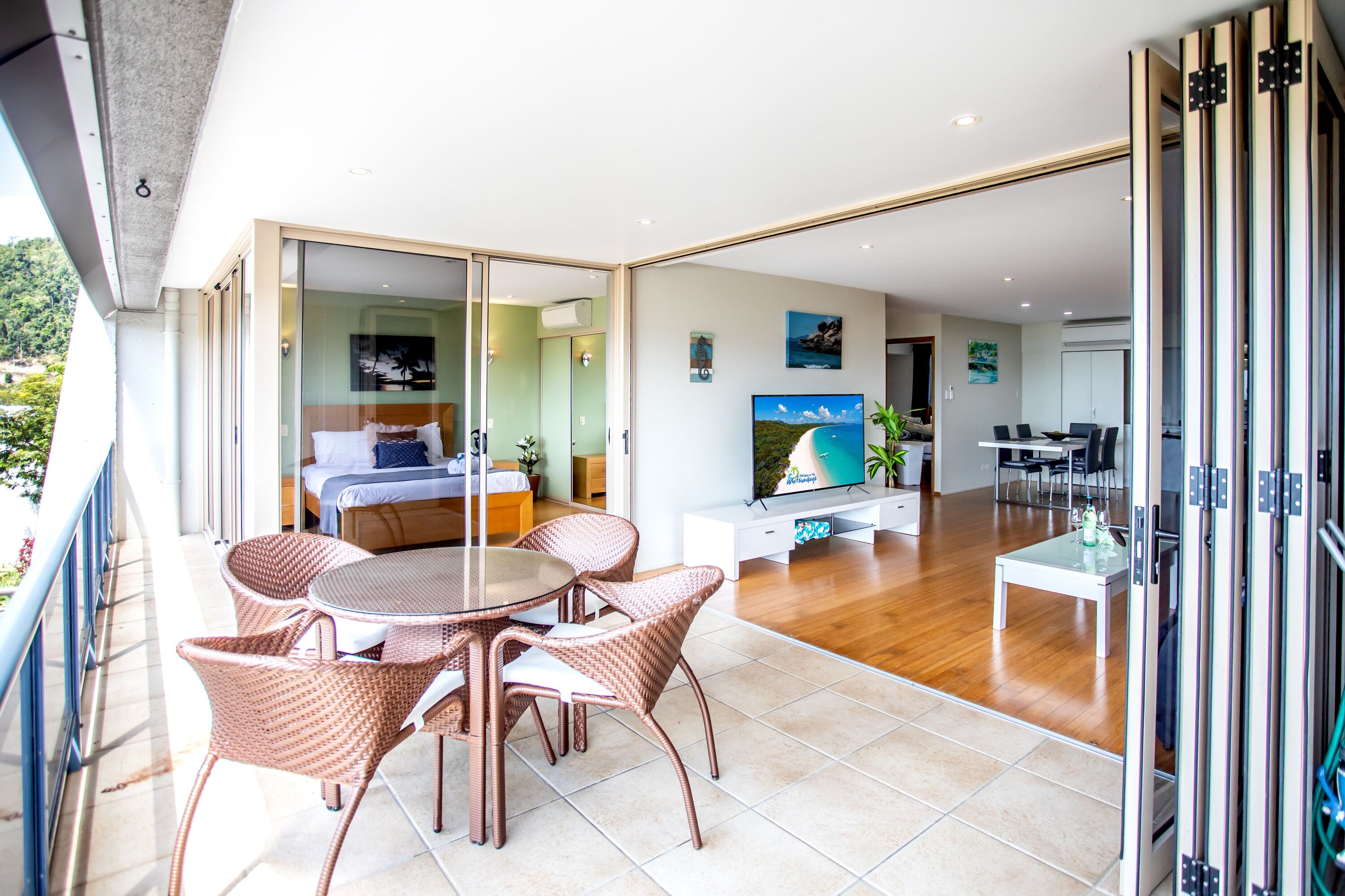 Un salon moderne comprenant un coin repas, un téléviseur et un balcon aménagé avec des meubles en osier.
