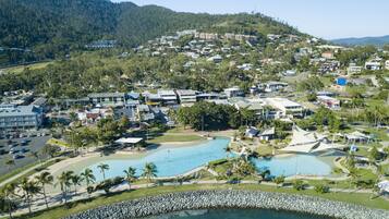An aerial view of a coastal resort with multiple swimming pools, a rocky shoreline, and a backdrop of a forested hill.