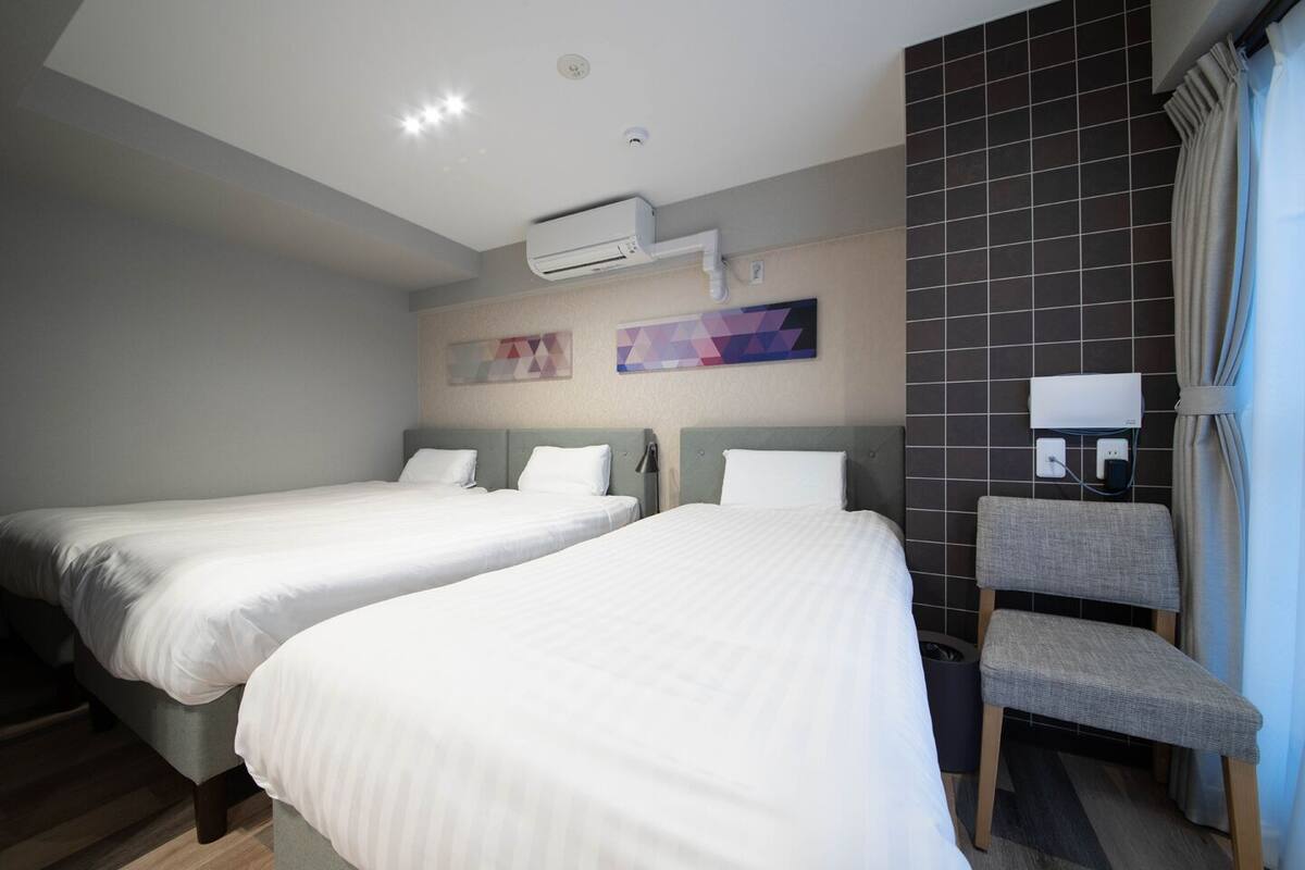 Quarto de hotel com duas camas, uma poltrona cinza estofada, ar condicionado fixado na parede e parede revestida de azulejos com uma obra de arte geométrica.