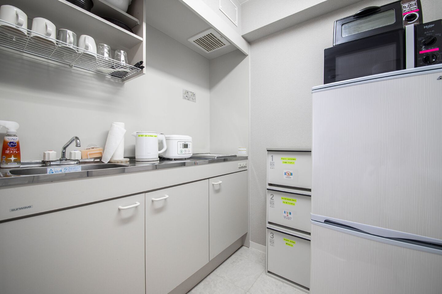Eine kleine Küche mit einer Mikrowelle, einer Kaffeemaschine und einem Kühlschrank. Es gibt Schränke und ein Regal mit Tassen.