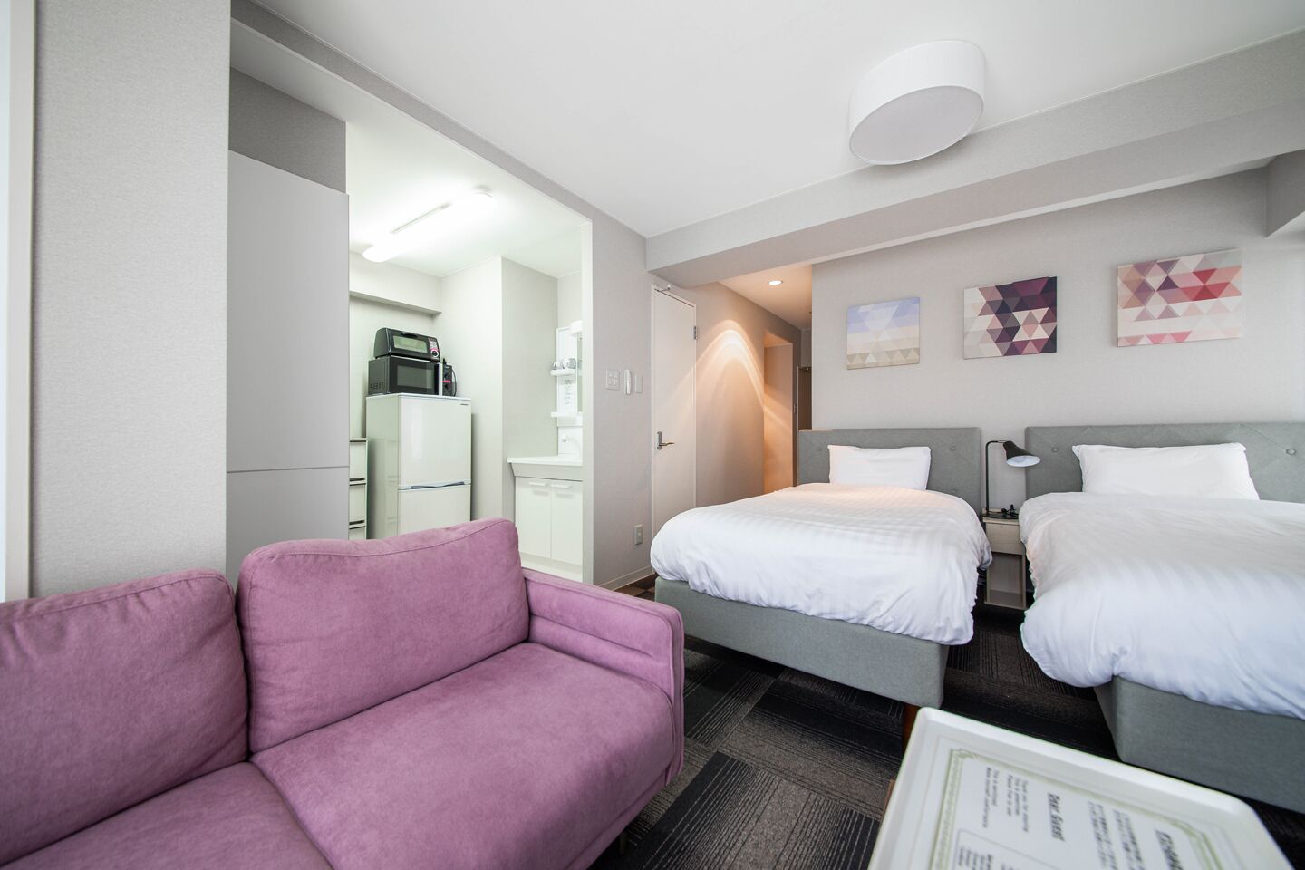 Quarto de hotel com uma cama cinza, um sofá rosa e um frigorífico branco.