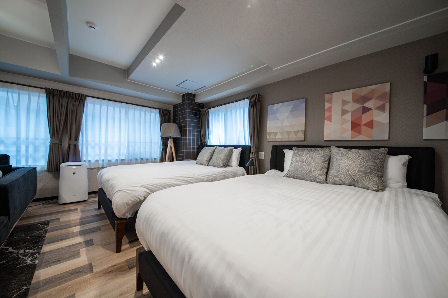 A modern hotel room with two large beds, a sofa, a lamp, and a view of the city.