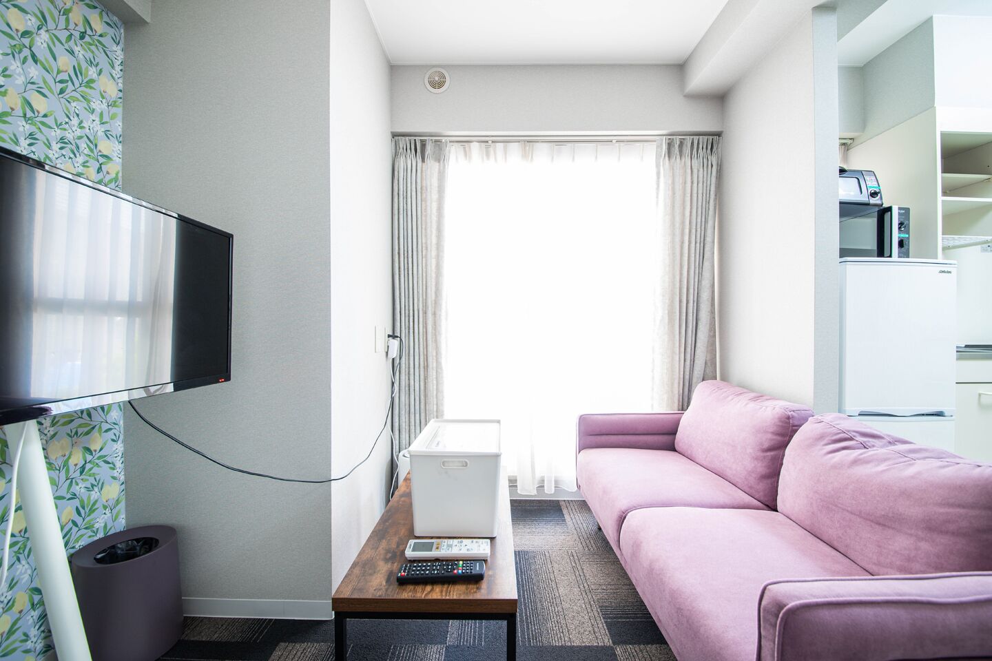 A hotel room with a flat-screen TV, a white mini-fridge, a pink sofa, a wooden coffee table, and a trash bin.