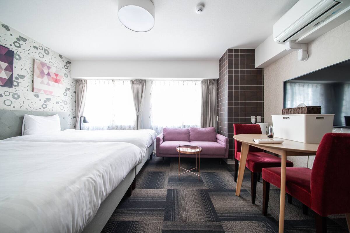 Quarto de hotel com duas camas, um sofá, uma mesa pequena, uma escrivaninha com cadeiras e um aparelho de ar condicionado.