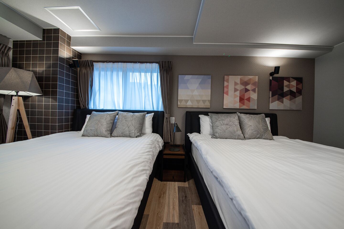 A hotel room with two beds, a window with curtains, a lamp, and wall art.